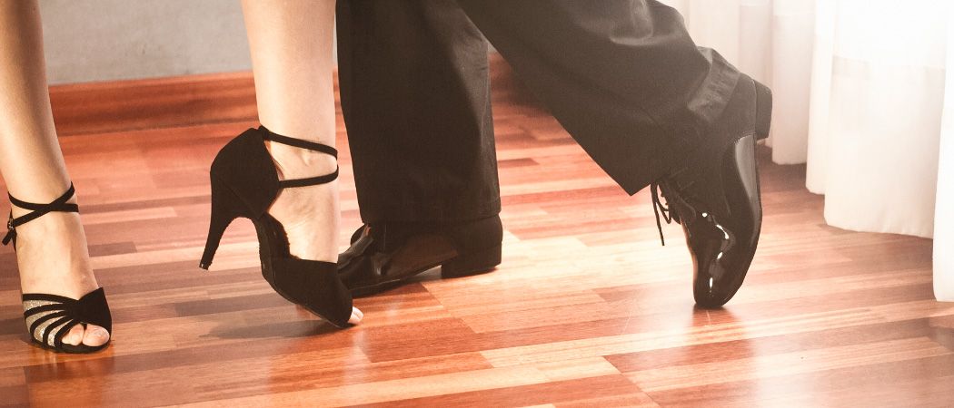 Ein Paar in formeller Kleidung tanzt eng auf einem Holzboden, beide tragen schwarze Schuhe mit sichtbaren Schnürsenkeln und Absätzen.