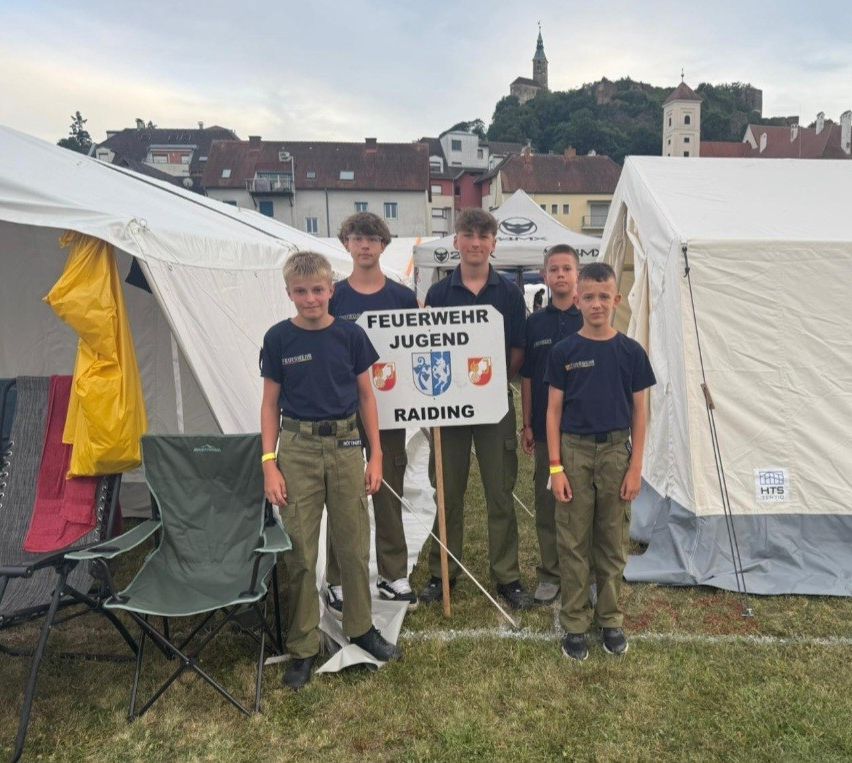 Fünf Jungen in Uniform posieren vor Zelten und halten ein Schild mit der Aufschrift 'Feuerwehr Jugend Raiding'. Dahinter ist ein Hügel mit Gebäuden und einem klaren Himmel zu sehen.