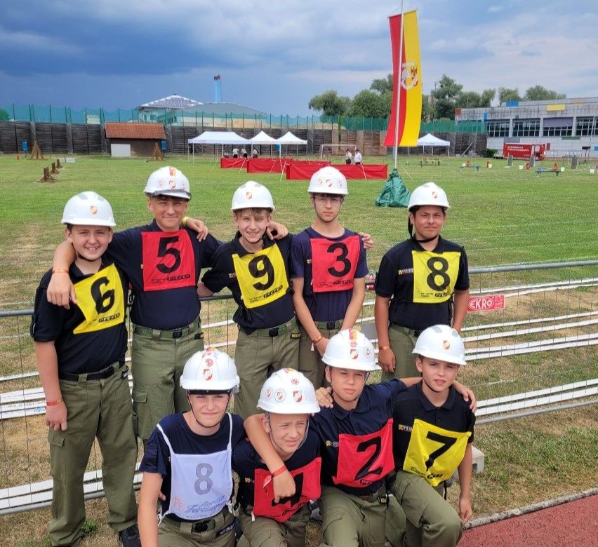 Eine Gruppe junger Jungen in Helmen und nummerierten Westen posiert für ein Foto auf einem Sportfeld mit Zelten und einem Gebäude im Hintergrund.