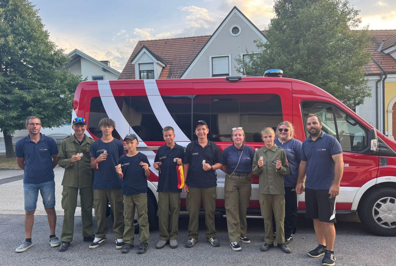 Eine Gruppe von Personen in blauen Uniformen und Mützen steht vor einem roten Van und posiert für ein Foto. Dahinter befinden sich Häuser und Bäume.