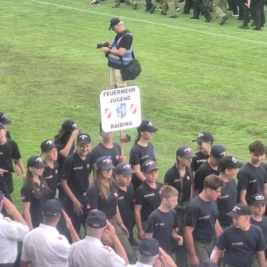 A group of young people in uniform, possibly a youth organization, stands on a field with a sign that reads 'Feuerwehr Jugend Raiding'. A man with a camera is standing behind them.