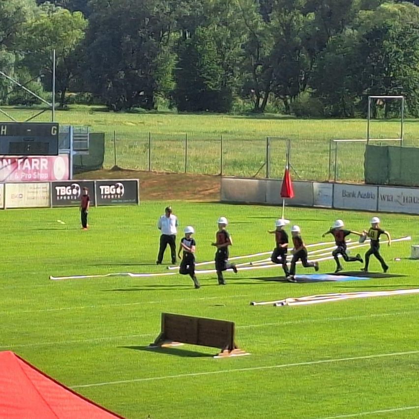 A group of children in helmets and sneakers are running on a field with a red flag in the background. There are adults standing on the sidelines.