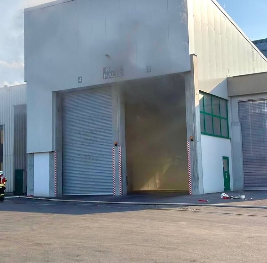 Ein Gebäude mit einer großen offenen Garagentür, die mit 'KAHLE HALL 4' beschriftet ist, mit Rauchaustritt und anwesenden Feuerwehrleuten.