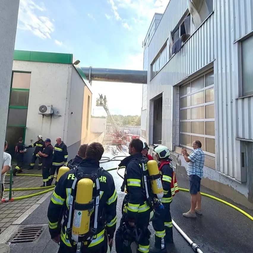 Feuerwehrleute stehen vor einem Gebäude mit einem Feuer, das durch ein zerbrochenes Fenster sichtbar ist. Sie sind mit Helmen und Sauerstoffflaschen ausgestattet.