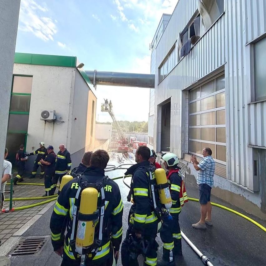 Feuerwehrleute stehen vor einem Gebäude mit einem Feuer, das durch ein zerbrochenes Fenster sichtbar ist. Sie sind mit Helmen und Sauerstoffflaschen ausgestattet.