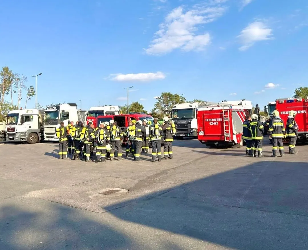 Eine Gruppe Feuerwehrleute in gelben und schwarzen Uniformen versammelt sich auf einem Parkplatz, mit Feuerwehrwagen im Hintergrund. Der Himmel ist blau mit einigen Wolken.