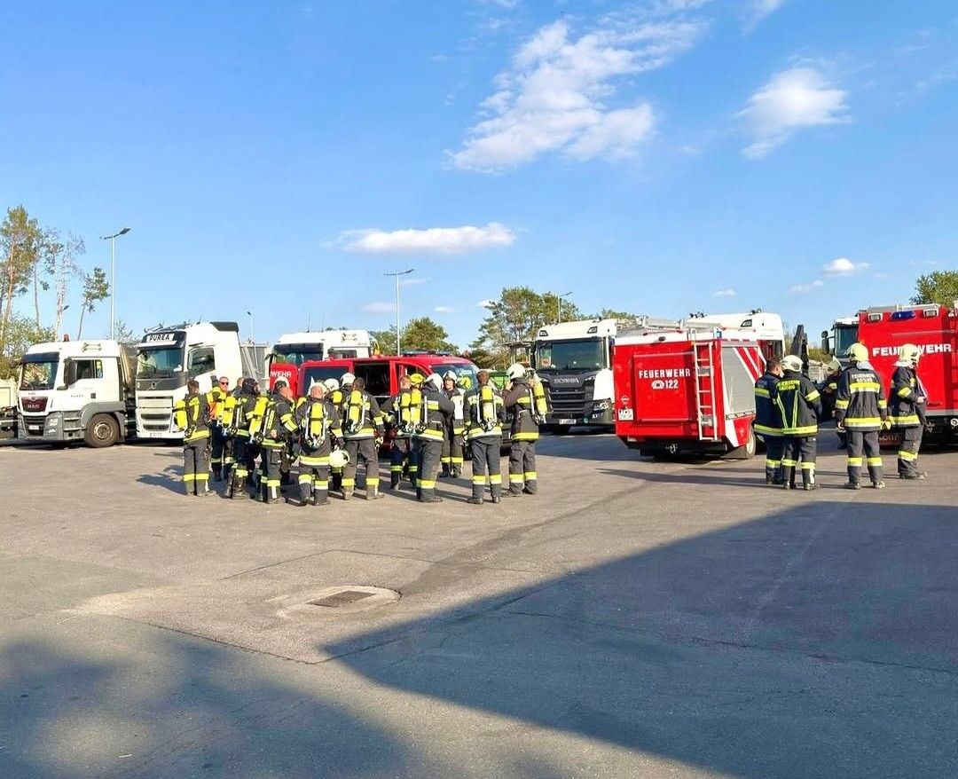 Eine Gruppe Feuerwehrleute in gelben und schwarzen Uniformen versammelt sich auf einem Parkplatz, mit Feuerwehrwagen im Hintergrund. Der Himmel ist blau mit einigen Wolken.