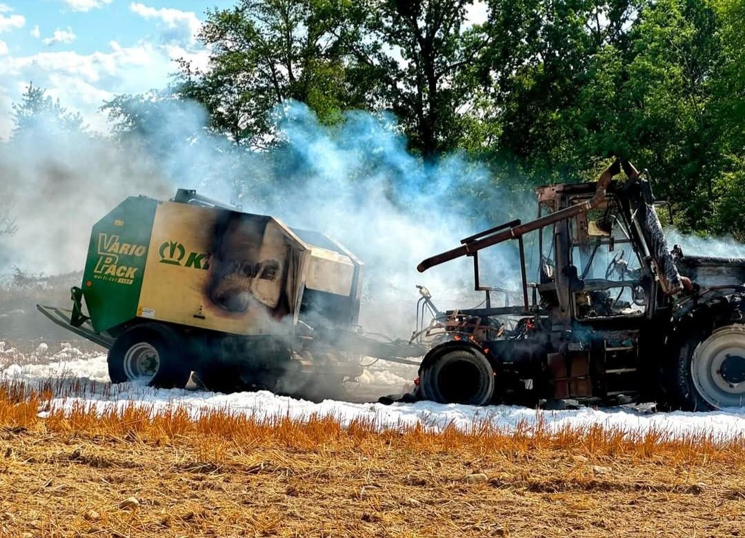 A burnt-out machinery vehicle with smoke billowing out, lying on a field with dry grass. The vehicle has a green and yellow color scheme.
