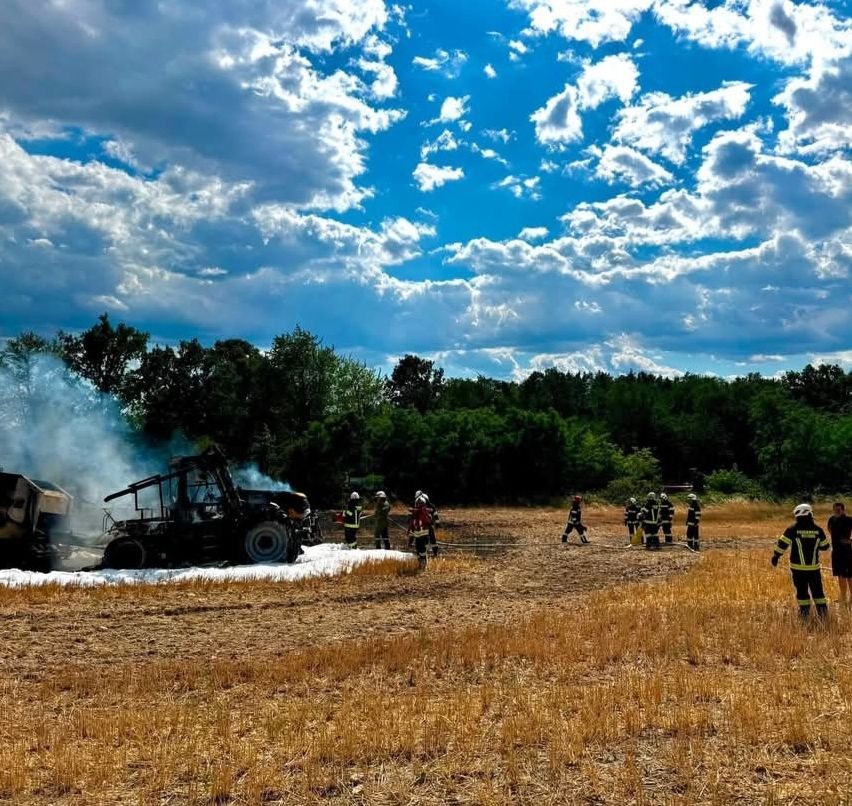 Firefighters in full gear are working to extinguish a burning tractor in a field with dry grass and a blue sky with clouds in the background.