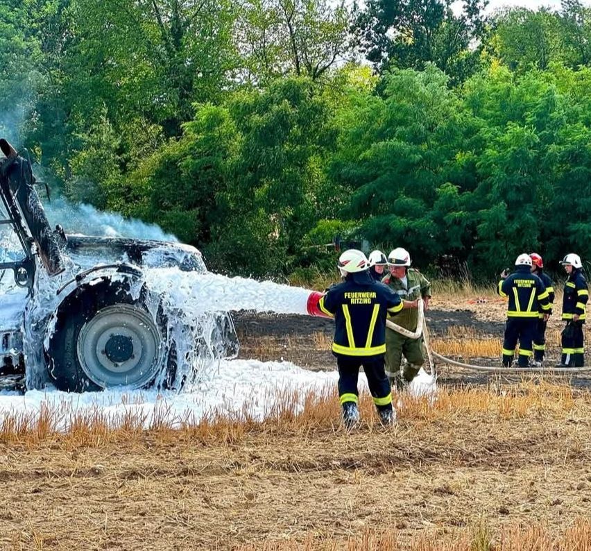 Firefighters in protective gear are using a hose to extinguish a burning vehicle in a field.