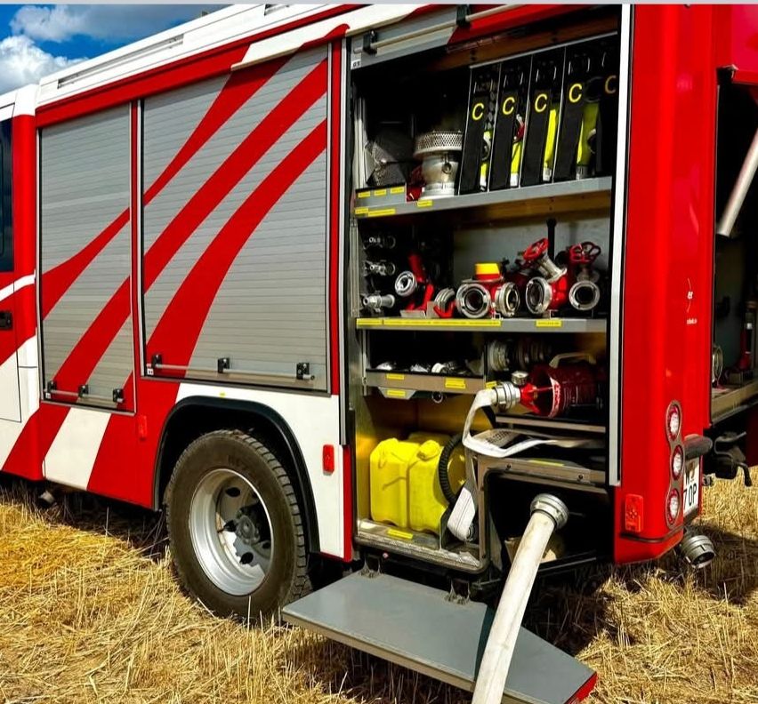 A red and white fire truck with its back doors open, revealing a well-organized array of fire-fighting equipment, including hoses, nozzles, and yellow containers, parked in a field.