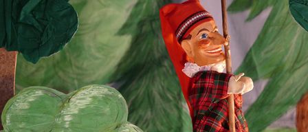 A puppet wearing a red hat and plaid clothing is climbing up a wooden stick in a forest setting.