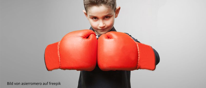 Ein junger Junge mit kurzen Haaren, der ein schwarzes Hemd trägt, hält rote Boxhandschuhe hoch. Er sieht entschlossen aus.