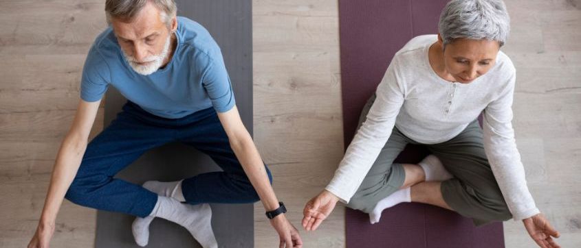 Zwei ältere Personen praktizieren Yoga auf Holzböden, jede mit einer Yogamatte, eine in Blau und die andere in Weiß.