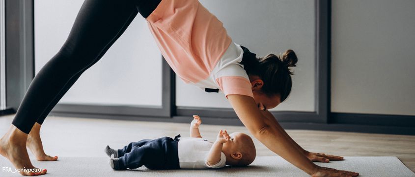 Eine Frau in Trainingskleidung macht Yoga-Posen mit einem Baby, das vor ihr auf dem Boden liegt.