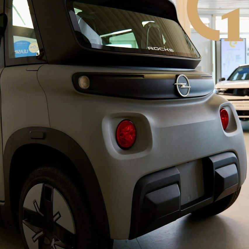 Ein grauer Opel Rocks Elektroauto steht in einer Ausstellung. Die Rückseite zeigt einen kleinen Spoiler, Rücklichter und einen integrierten Nummernschild im hinteren Stoßfänger.