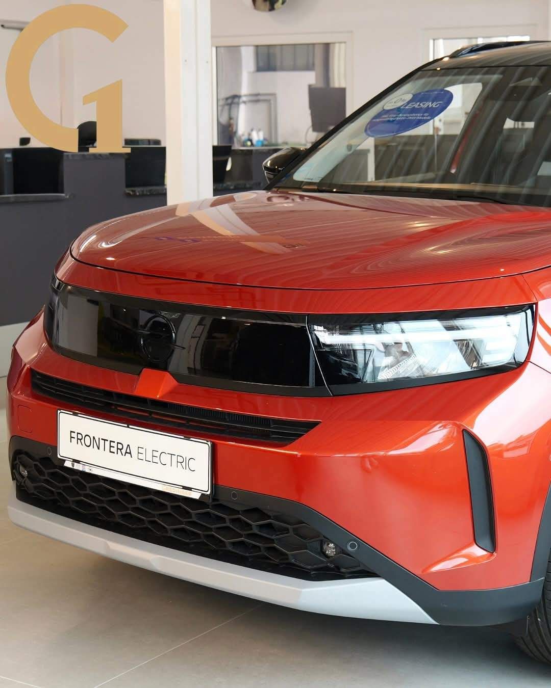 Ein roter Frontera Electric-Wagen wird in einem Ausstellungsraum präsentiert, wobei der schlanke Stoßfänger vorne und die Scheinwerfer hervorgehoben werden, mit einem Leasing-Schild auf der Windschutzscheibe.