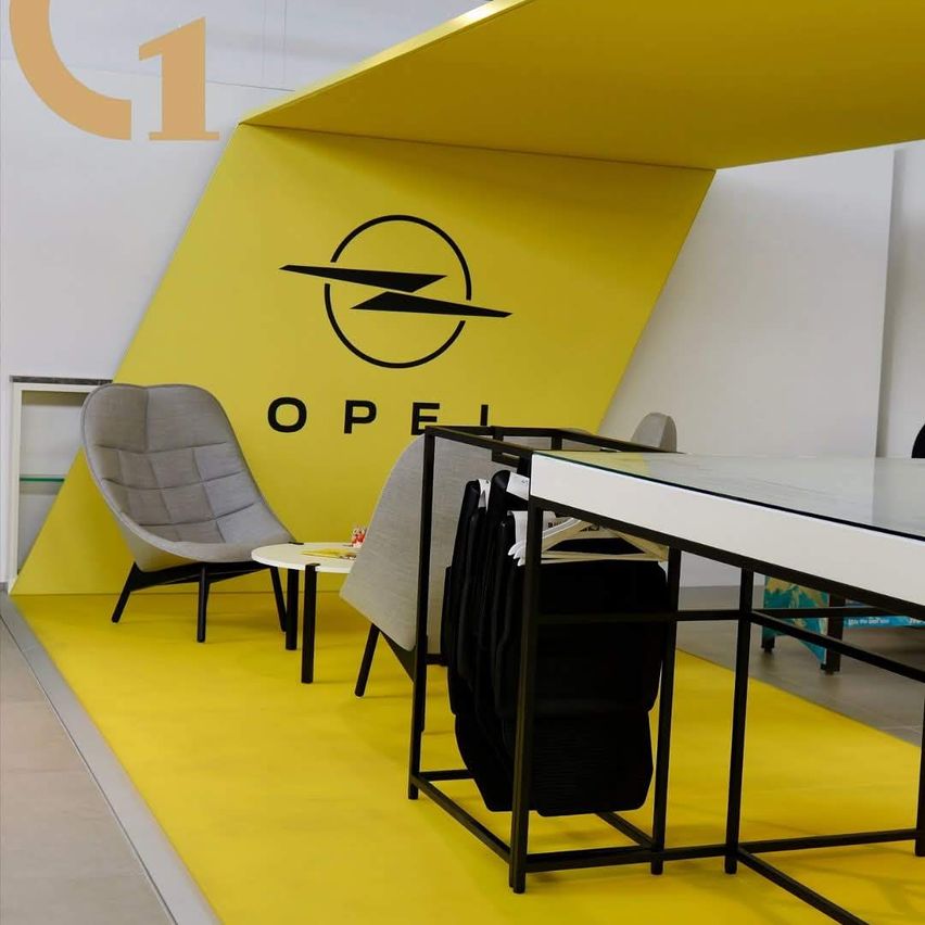 Ein Opel-thematischer Raum mit gelbem Boden, gelber Wand mit Opel-Logo und grauen Stühlen. Ein Schreibtisch mit hängenden Kleidern und ein Tisch in der Nähe.