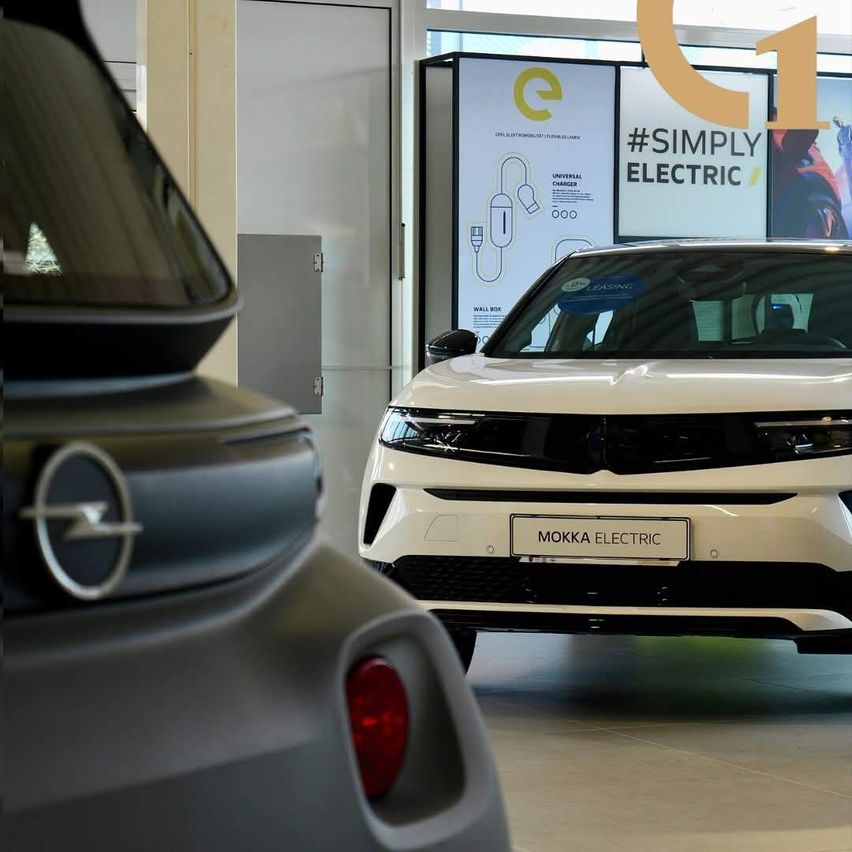 Zwei Elektroautos sind in einer Ausstellungshalle ausgestellt. Das Auto auf der linken Seite hat ein Opel-Logo und das Auto auf der rechten Seite ist ein Mokka Electric. Die Anzeige dahinter hat ein gelbes und schwarzes Thema mit einem Ladesymbol.