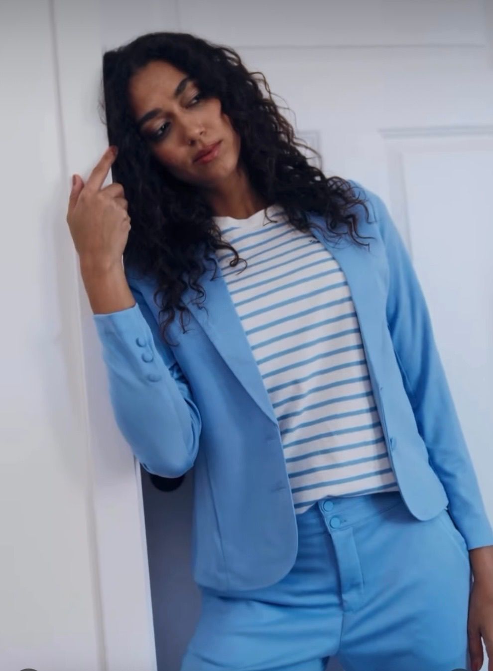 Eine Frau mit lockigem Haar, die einen hellblauen Blazer über einem weiß-blau gestreiften Shirt trägt, lehnt sich gegen eine weiße Tür.