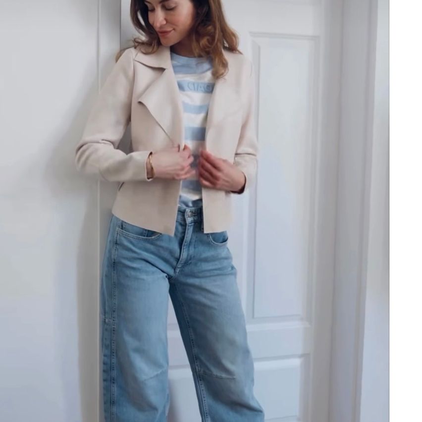 Eine Frau steht vor einer weißen Tür, trägt eine beige Jacke und hellblaue Jeans und lächelt, während sie ihre Jacke justiert.