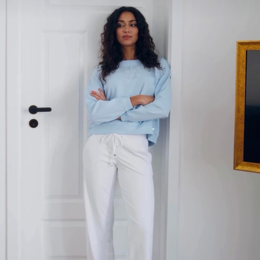 Eine Frau steht vor einer weißen Tür, trägt ein hellblaues Sweatshirt und weiße Hosen, hat die Arme verschränkt und sieht ernst aus.