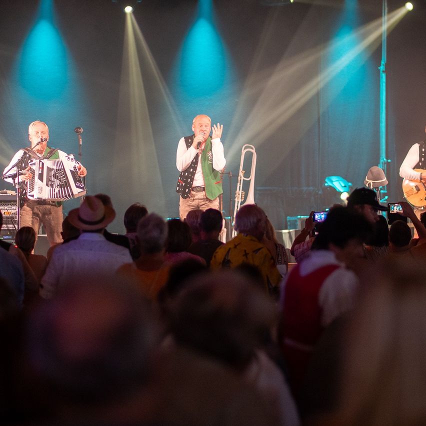 Eine Band tritt auf der Bühne vor einem Publikum auf. Der Leadsänger hält ein Mikrofon, ein anderer spielt Akkordeon und ein Dritter spielt Posaune. Scheinwerfer und blaue Lichter beleuchten die Bühne.
