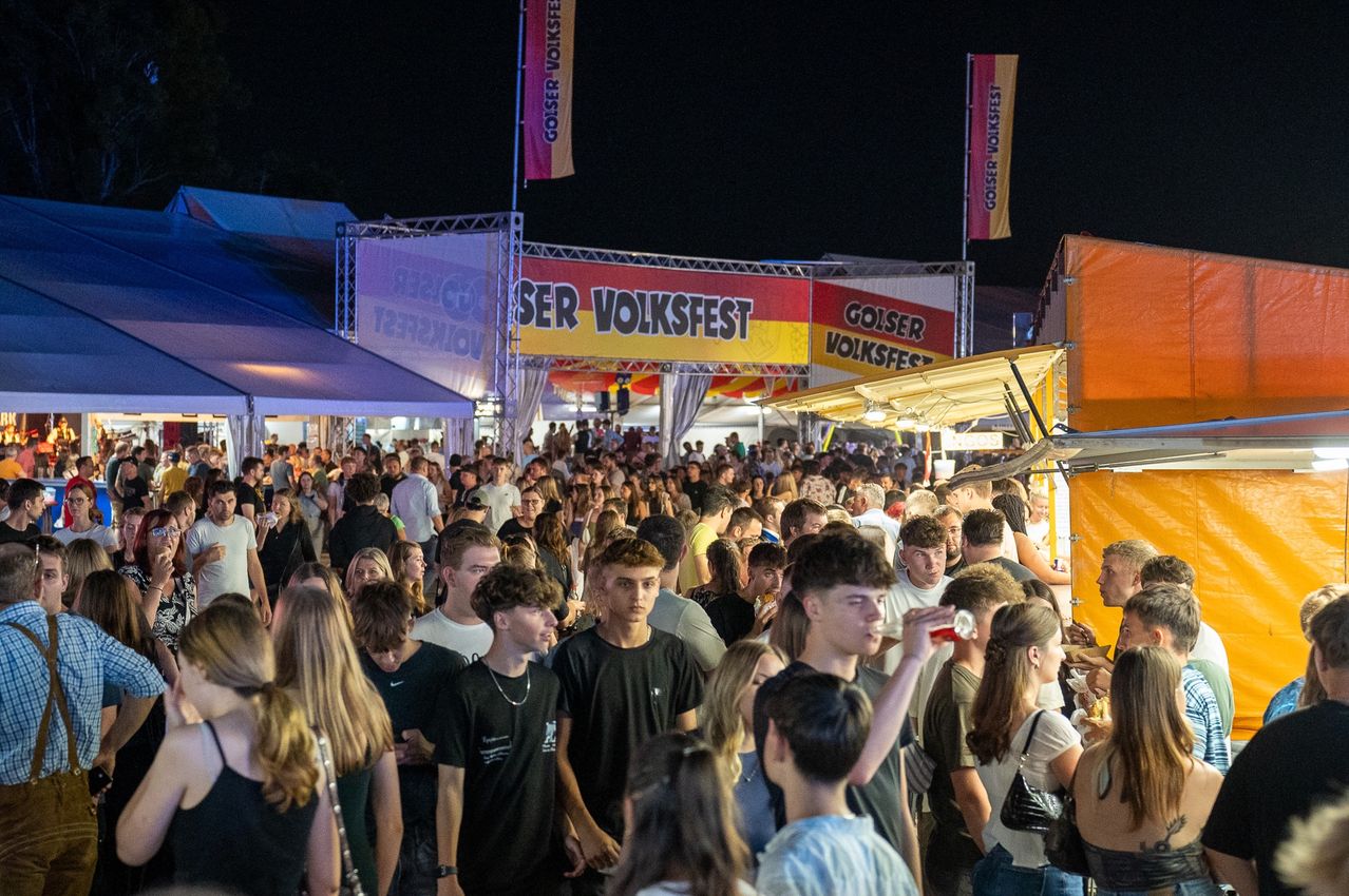Eine große Menschenmenge auf einem Festival bei Nacht, Banner oben, eines mit der Aufschrift 'GOSER VOLKSFEST', viele Teilnehmer in schwarzen Shirts, einige trinken aus Dosen.