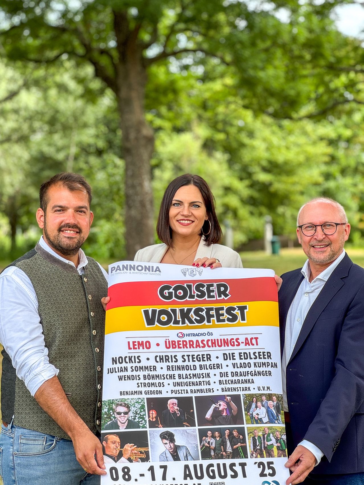 Drei Personen stehen in einem Park und halten ein großes Plakat, das das Golser Volksfest mit Bildern von Künstlern bewirbt. Der Mann auf der linken Seite trägt eine Weste, die Frau ein weißes Oberteil und der Mann auf der rechten Seite eine Brille. Bäume und Gras sind im Hintergrund.