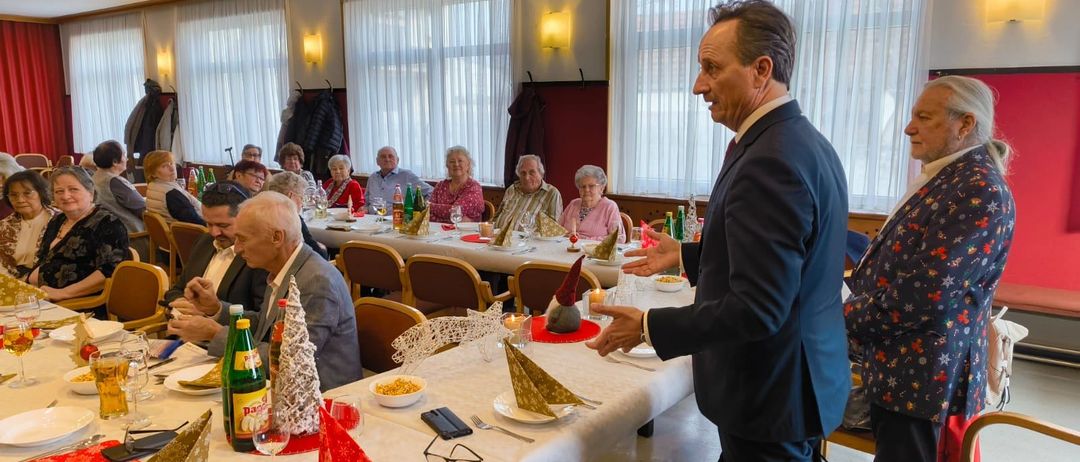 Ein Mann in einem Anzug spricht zu einem Tisch voller älterer Menschen. Der Tisch hat einen Weihnachtsbaum, eine Kerze und verschiedene Gerichte. Der Raum hat weiße Vorhänge und Lampen.