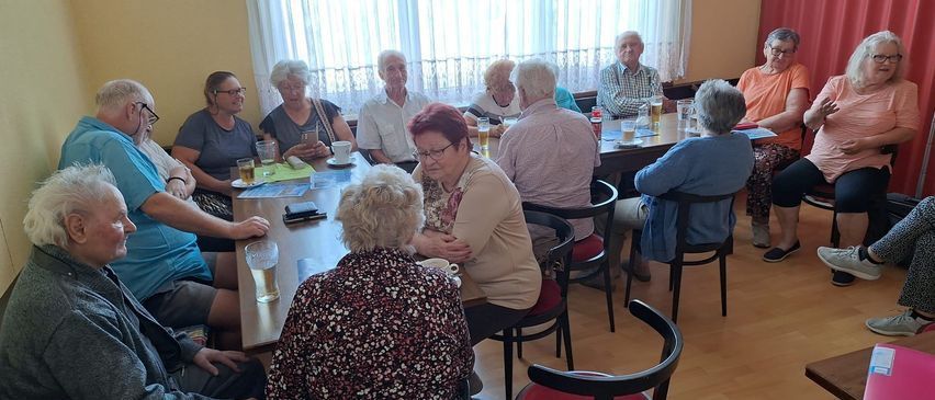 Eine Gruppe älterer Erwachsener sitzt in einem Raum um einen Tisch, einige unterhalten sich. Auf dem Tisch stehen Gläser mit Bier und Tassen.