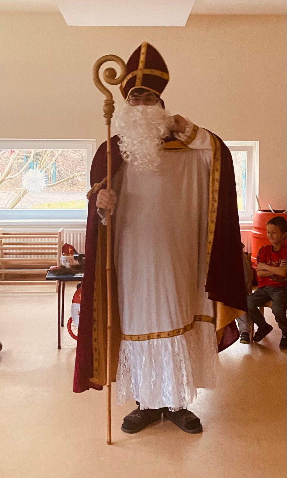 Eine Person in einer Weihnachtsmann-Kostümierung steht mit einem Stock in einem Raum. Ein Junge sitzt auf einem Stuhl neben ihm. In der Nähe steht ein Tisch mit einem Weihnachtsmannhut.