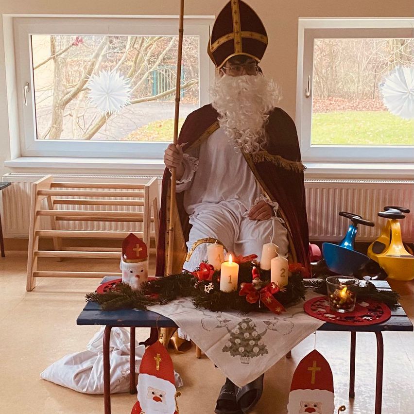 Eine Person in der Kleidung des Heiligen Nikolaus hält einen Stab und sitzt an einem Tisch mit einer Adventskrone und Kerzen. Die Umgebung ist innen mit Fenstern, die Bäume draußen zeigen.