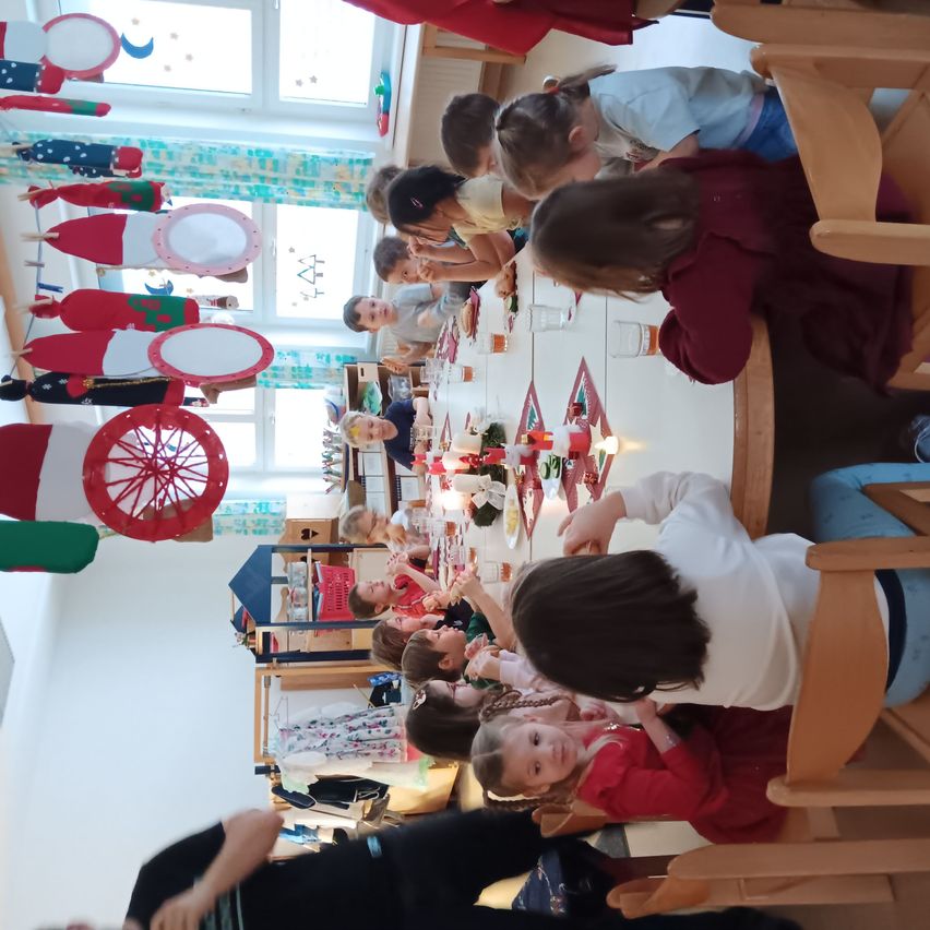 Kinder in einem Klassenzimmer sitzen um einen Tisch herum, essen und spielen. Der Raum ist mit Weihnachtsdekorationen geschmückt. Eine Person steht links.