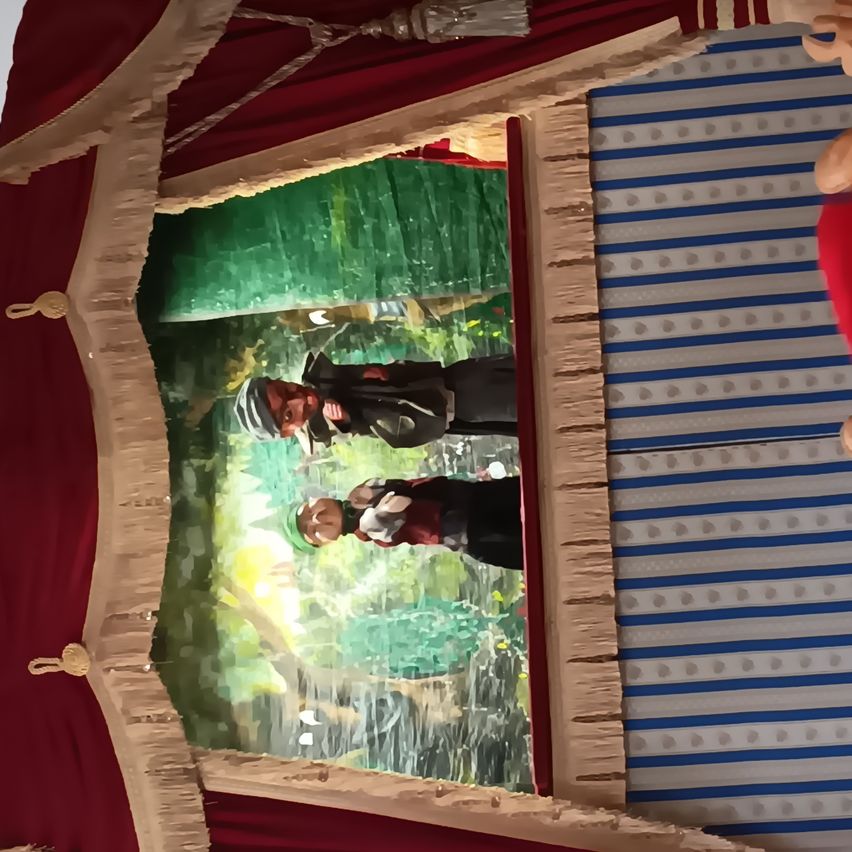 Eine Draufsicht auf eine kleine Bühneneinrichtung mit rotem Vorhang als Hintergrund. Zwei Figuren stehen auf der Bühne, eine kniend und eine stehend, mit grünem Hintergrund.