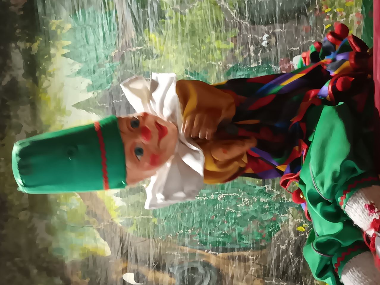 Eine bemalte Puppe mit grünem Hut und bunter Kleidung steht unter einem Wasserfall. Der Hintergrund ist verschwommen mit grünem Laub.