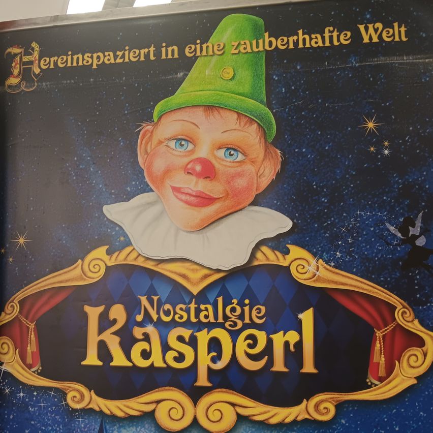 Ein Plakat zeigt einen fröhlichen Clown mit einem grünen Hut und einer roten Nase. Der Schriftzug 'Nostalgie Kasperl' steht in Gelb und ist von einem goldenen Rahmen umgeben. Der Hintergrund zeigt einen Sternenhimmel voller Wunder.