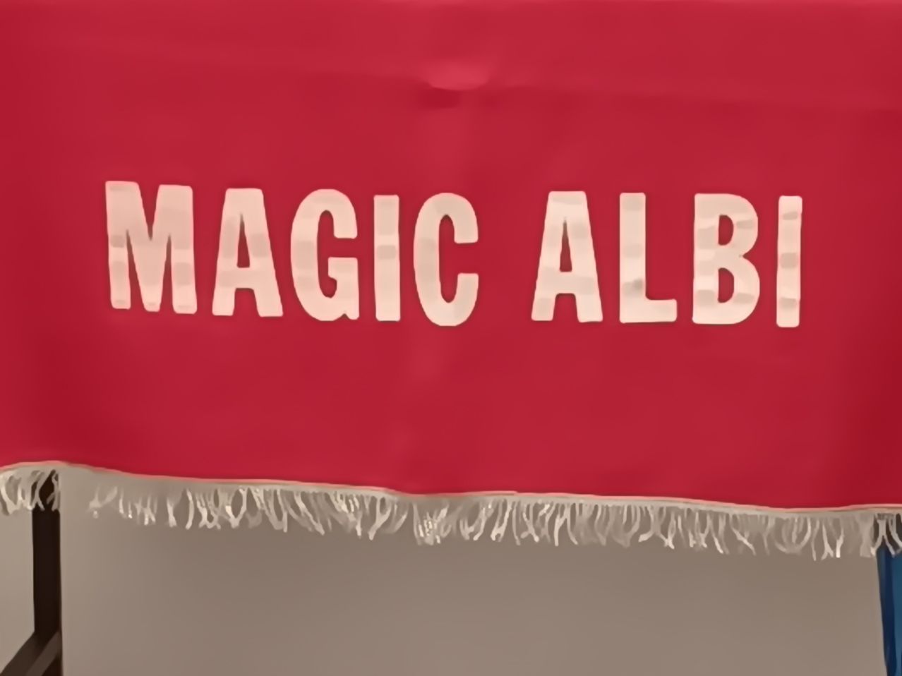 Eine Nahaufnahme eines roten Banners mit weißen Fransenkanten, auf dem die Worte 'MAGIC ALB' in großen weißen Buchstaben stehen.