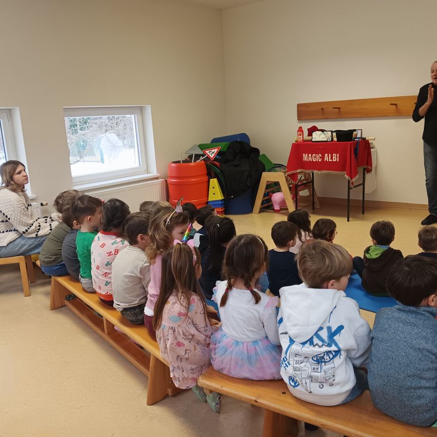 Ein Klassenzimmer mit einer Gruppe von Kindern, die auf Bänken sitzen. Ein Mann steht vor einem Tisch mit einem roten Tuch, auf dem 'MAGIC ALB' steht. Hinter dem Mann befindet sich ein Fenster und in der Nähe des Tisches steht ein Fass.