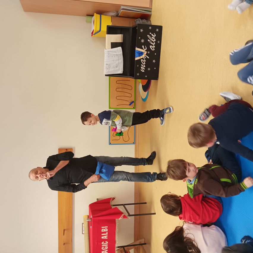 Ein Mann führt in einem Raum Zaubertricks für eine Gruppe von Kindern vor. Die Kinder beobachten ihn aufmerksam und sitzen auf einer blauen Matte. Es gibt eine schwarze Box mit den Worten 'Zauberei alles'.