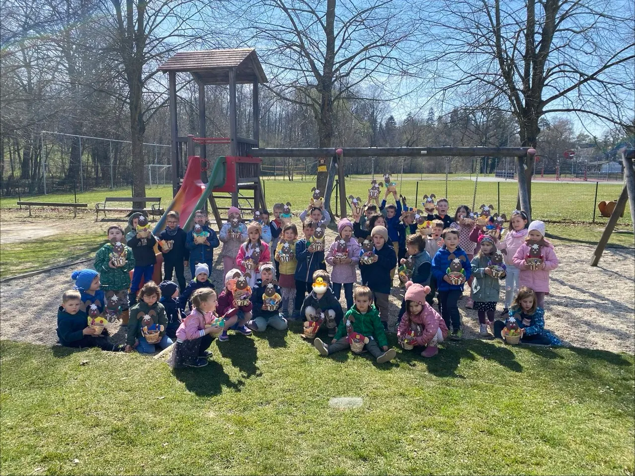 Eine Gruppe von Kindern posiert für ein Foto auf einem Rasenfeld und hält alle dekorierte Körbe. Ein Spielplatz mit Rutschen und Schaukeln ist im Hintergrund.