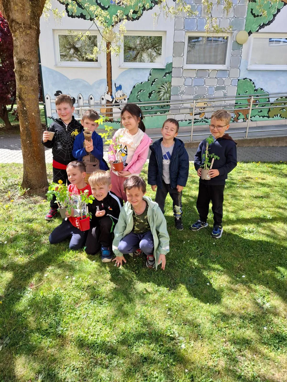 Kinder posieren für ein Bild draußen mit Topfpflanzen in den Händen, ein Gebäude mit Wandgemälden im Hintergrund.