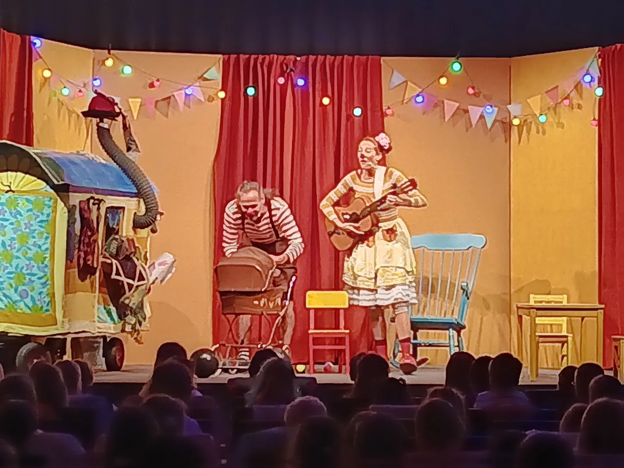 Auf einer Bühne führen zwei Personen mit farbenfrohem Hintergrund auf. Eine Person spielt Gitarre, die andere beugt sich vor. Stühle und ein Kinderwagen sind vorhanden. Ein Publikum schaut zu.