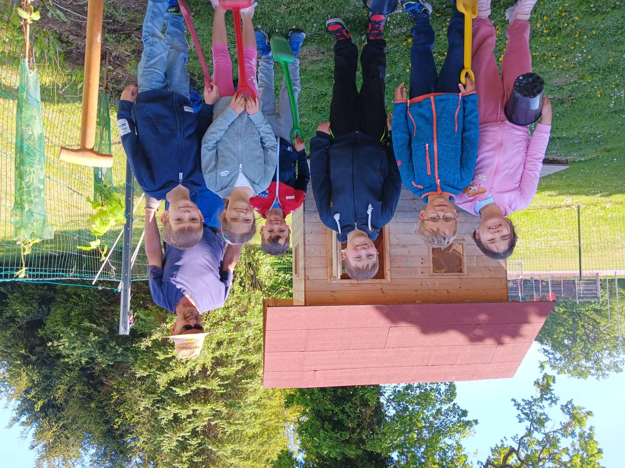 Gruppe von Kindern, die auf den Kopf stehend um ein kleines Holzhaus mit Werkzeugen auf einer Rasenfläche stehen.