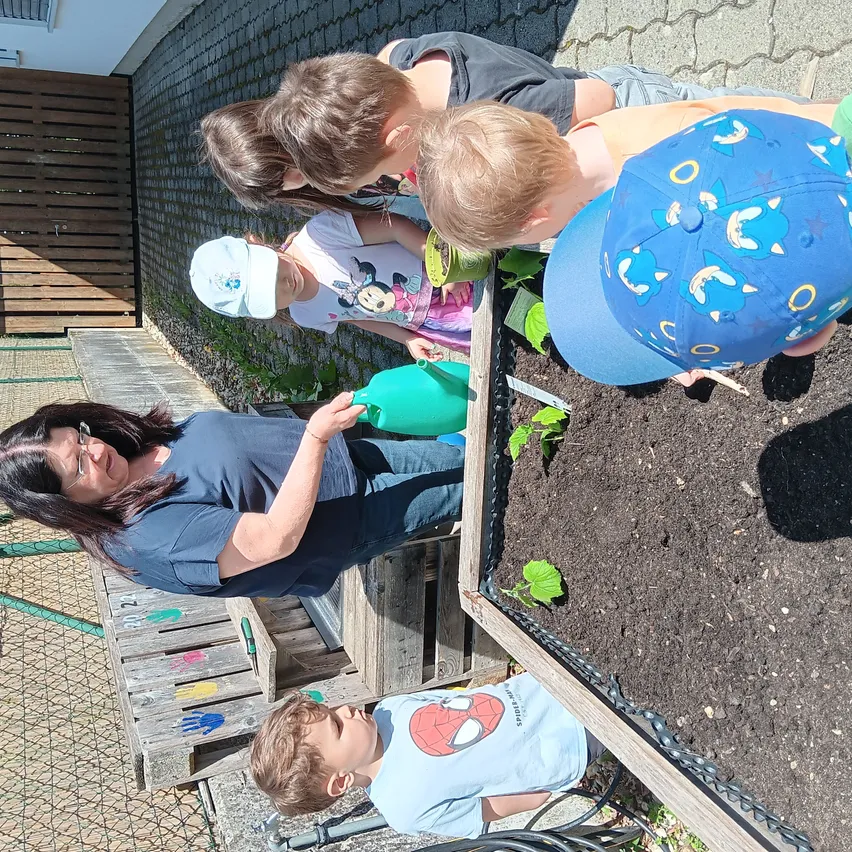 Eine Frau und Kinder pflanzen im Garten. Ein Kind gießt Pflanzen, andere schauen sich die Pflanzen genau an. Die Frau beugt sich über ein Hochbeet.