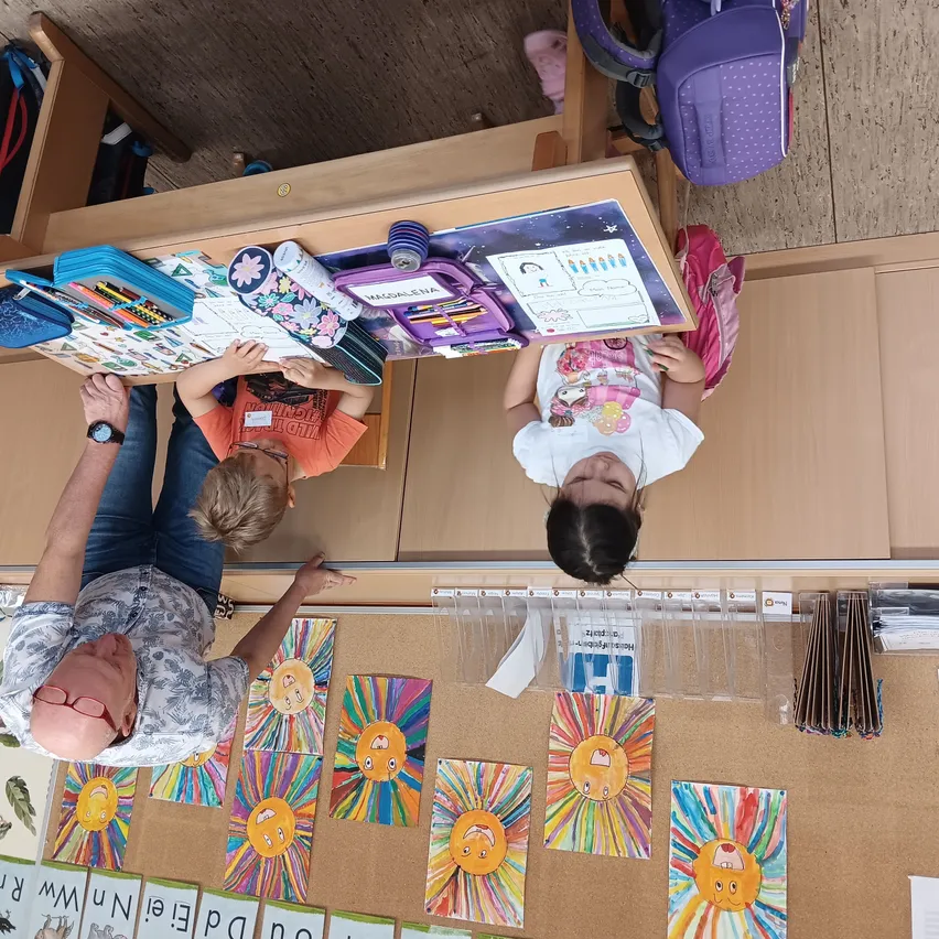 Ein Mann und zwei Kinder betrachten Kunstwerke auf einem Tisch. Der Mann beugt sich nach unten, ein Kind schaut in ein Buch und das andere in ein Papier.