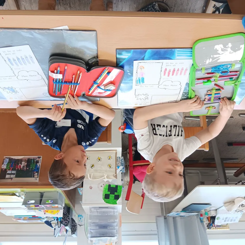Zwei Kinder lehnen sich über einen Tisch und greifen nach Gegenständen in einem Stiftetui und halten Buntstifte. Der Tisch hat Papiere mit Zeichnungen und Text. Der Junge links trägt ein blaues Shirt mit weißen Punkten, während der Junge rechts ein weißes Shirt mit schwarzem Text trägt.