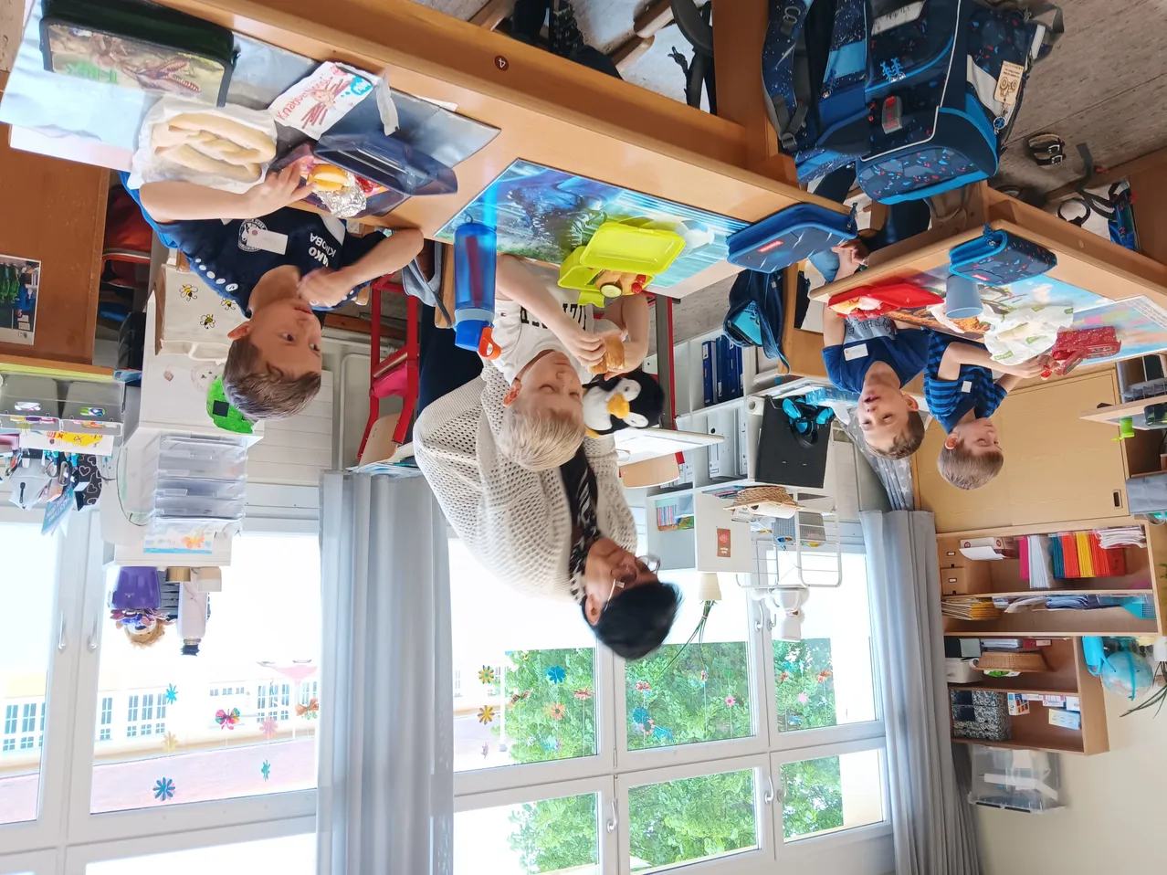 Eine Gruppe von Kindern sitzt an Tischen in einem Klassenzimmer. Einer isst, ein anderer blickt in ein Buch, und andere arbeiten. Eine Lehrerin lehnt sich über den Tisch und schaut nach unten.