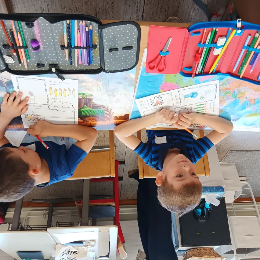 Zwei junge Jungen sitzen an einem Schreibtisch mit einer Karte. Sie zeichnen und malen mit Buntstiften. Eine Stifteetui und eine Schere sind in der Nähe.