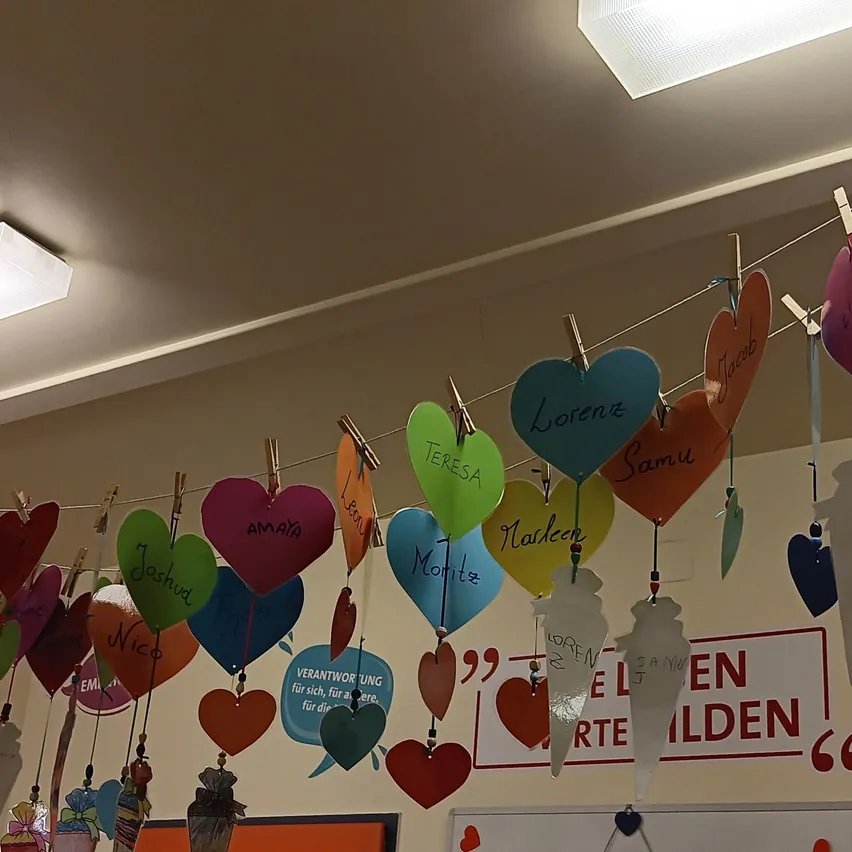 Eine Sammlung bunter herzförmiger Dekorationen mit Namen hängt an einer Schnur an einer Wand. Die Wand hat ein weißes Brett und ein rotes Schild mit weißen Buchstaben.
